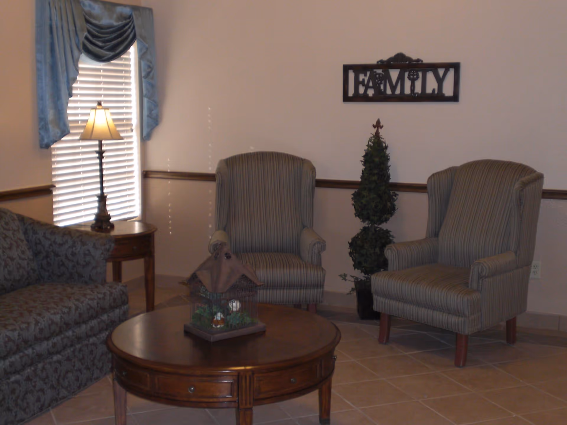 A living room seating area with two striped armchairs, a patterned sofa, round wooden coffee table, lamp by a window, and a 'FAMILY' wall sign.