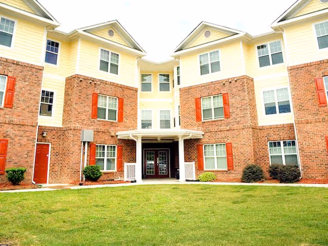 Three-story brick-and-siding apartment building with red shutters and a covered main entrance facing a grassy lawn.