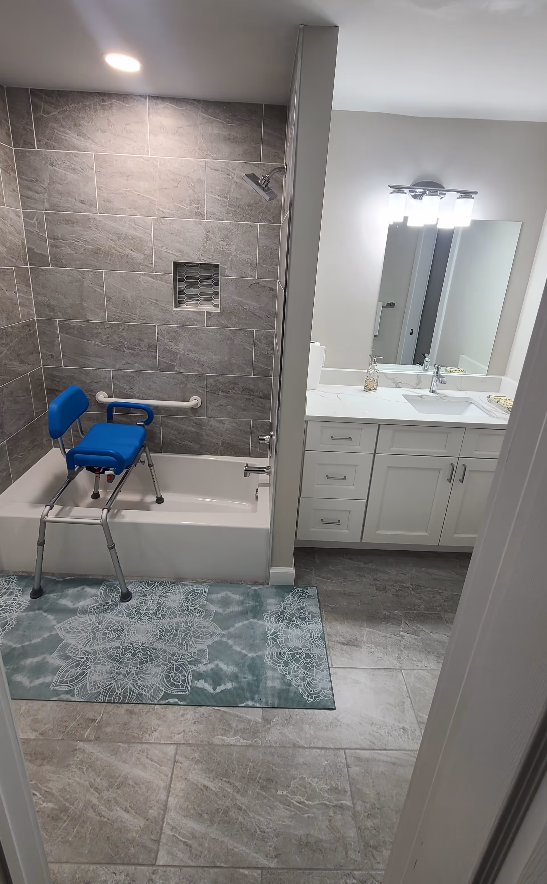 Bathroom with a bathtub featuring a blue shower chair and grab bar on the tiled wall. The floor has a patterned rug in front of the tub. To the right is a white vanity with a sink, a large mirror, and three light fixtures above it.