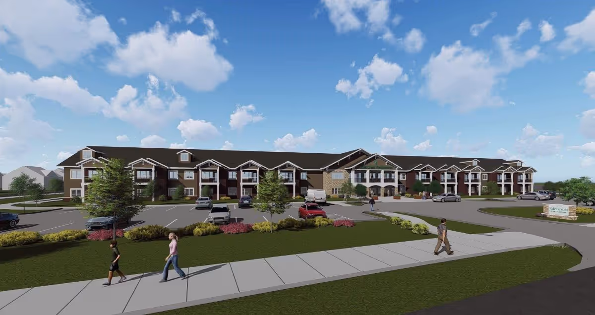 Exterior view of a large two-story senior living facility building with multiple balconies and a parking lot in front. There are a few people walking on the sidewalk and several cars parked. The sky is blue with scattered clouds.