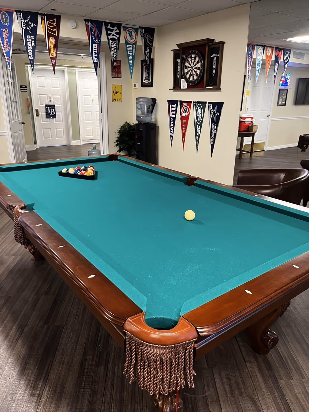 A pool table in a community game room decorated with sports pennants, a dartboard, and seating.