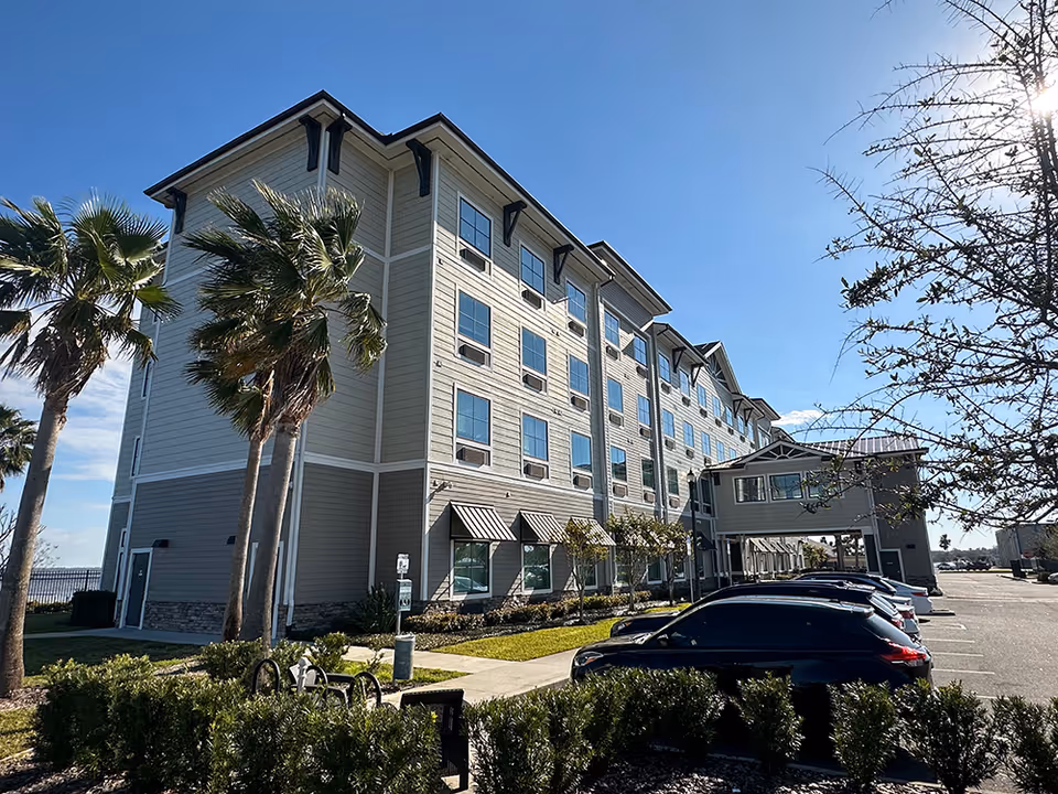 Exterior view of a multi-story assisted living facility building with several windows, palm trees, and parked cars in the parking lot under a clear blue sky.