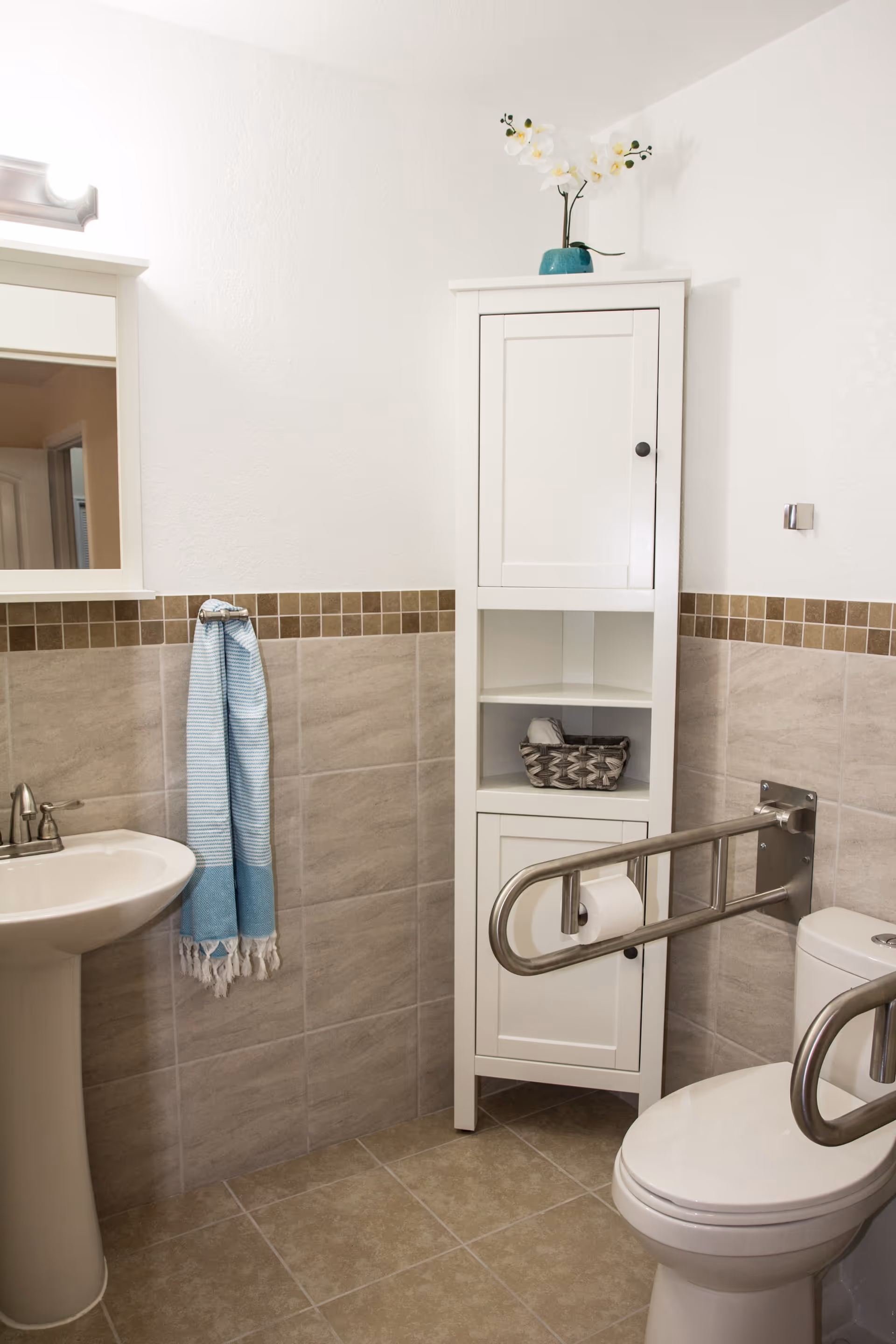 A clean and accessible bathroom featuring a white pedestal sink with a blue hand towel hanging on the wall, a white storage cabinet with a decorative plant on top, and a toilet equipped with stainless steel grab bars for support. The walls are tiled halfway up with beige tiles and a brown mosaic border, and the floor is tiled in a matching beige color.