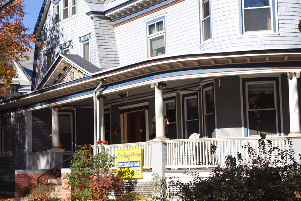 Exterior view of a large, gray, multi-story house with white trim and a wraparound porch. The porch has white railings and columns, with some plants and chairs visible. A yellow sign on the porch reads 'Sterling House Residential Care'. Trees with autumn-colored leaves are visible around the house.