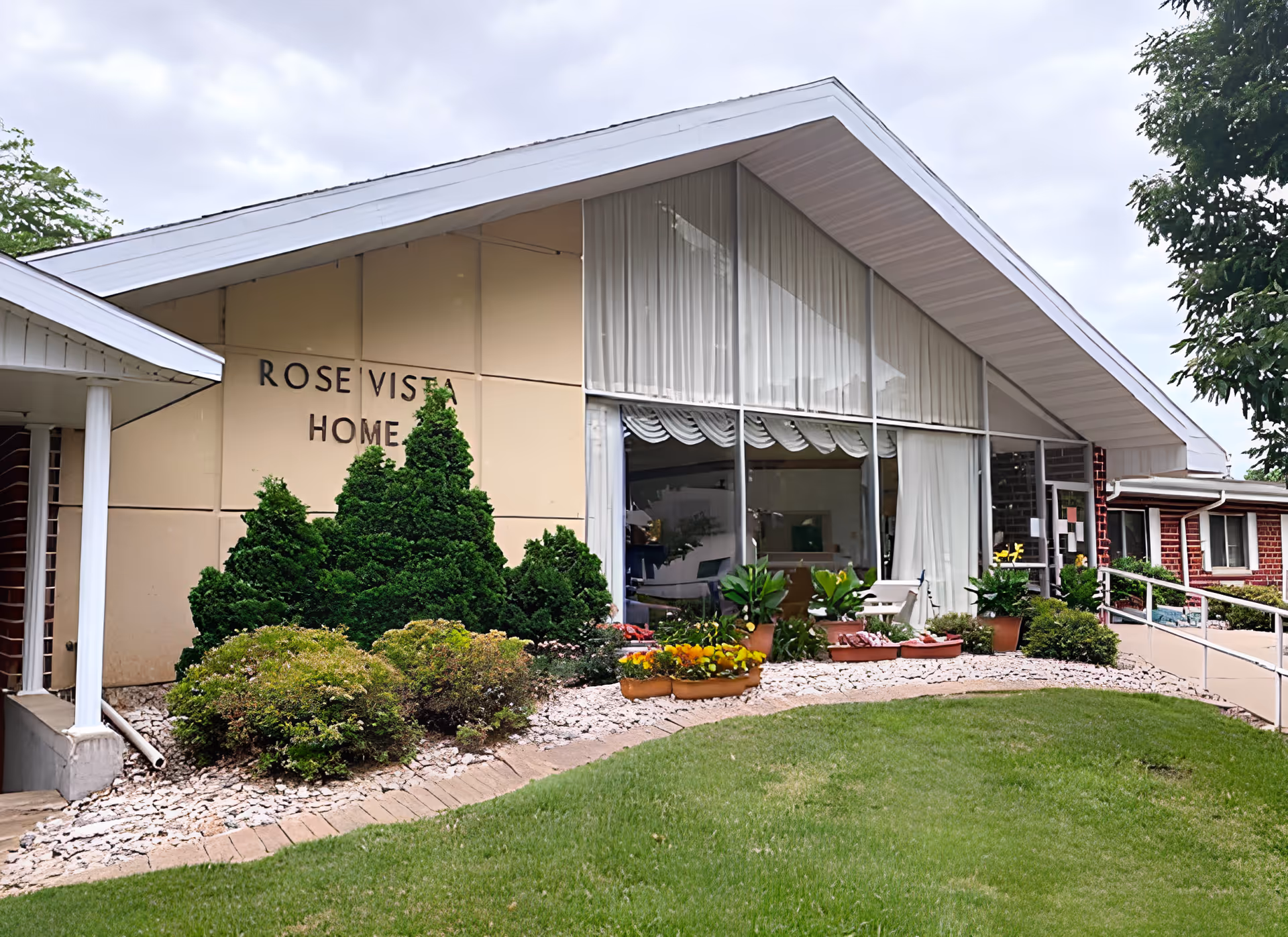 Front entrance of the Rose Vista Home building with large sloped roof, floor-to-ceiling windows, landscaping and potted plants.