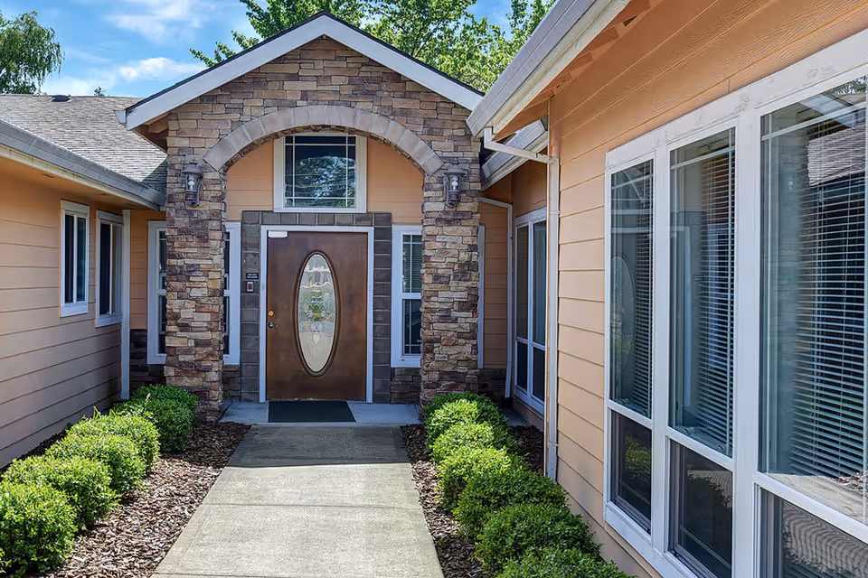 Entrance to a residential care facility with a stone archway surrounding a wooden door with an oval glass panel. The building has peach-colored siding, multiple windows with white frames, and neatly trimmed green bushes lining the concrete walkway leading to the door.