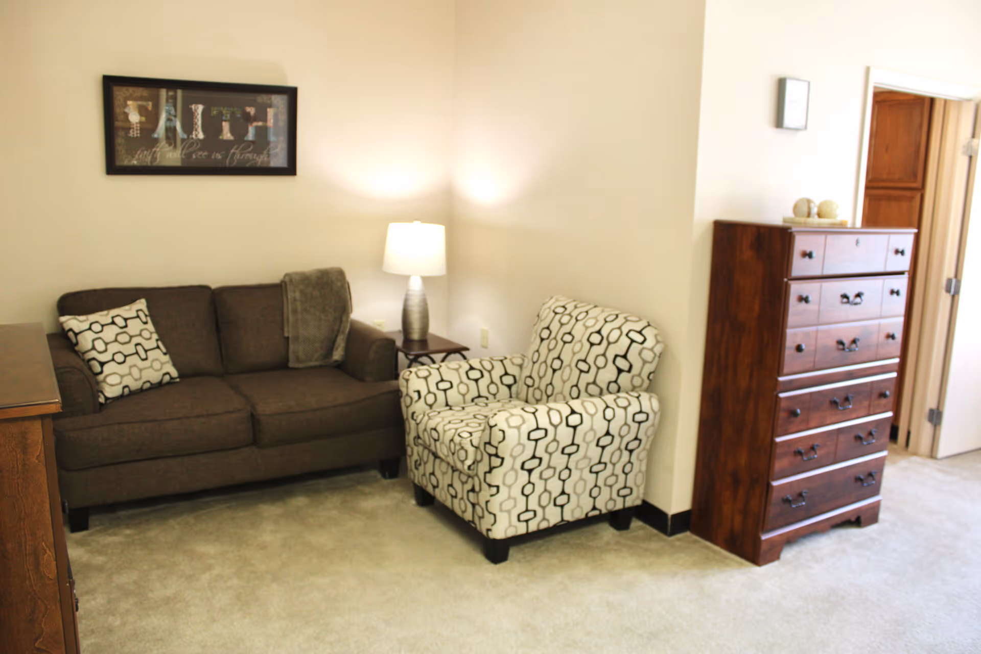 A cozy living room area with a brown sofa adorned with a patterned pillow and a gray throw blanket, a patterned armchair, a wooden chest of drawers, a small side table with a lamp, and a framed artwork on the wall that reads 'FAITH'.