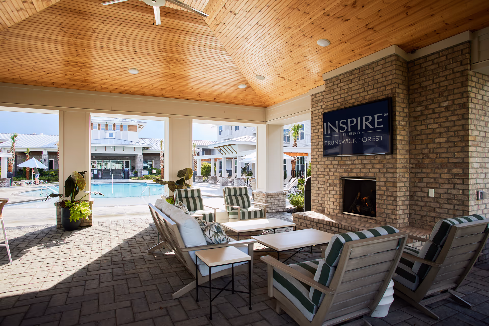 Covered outdoor seating area with cushioned chairs and sofas arranged around a fireplace. The fireplace has a mounted screen displaying the text 'INSPIRE BY LIBERTY BRUNSWICK FOREST'. Beyond the seating area, there is a swimming pool with lounge chairs and umbrellas, surrounded by buildings and palm trees.