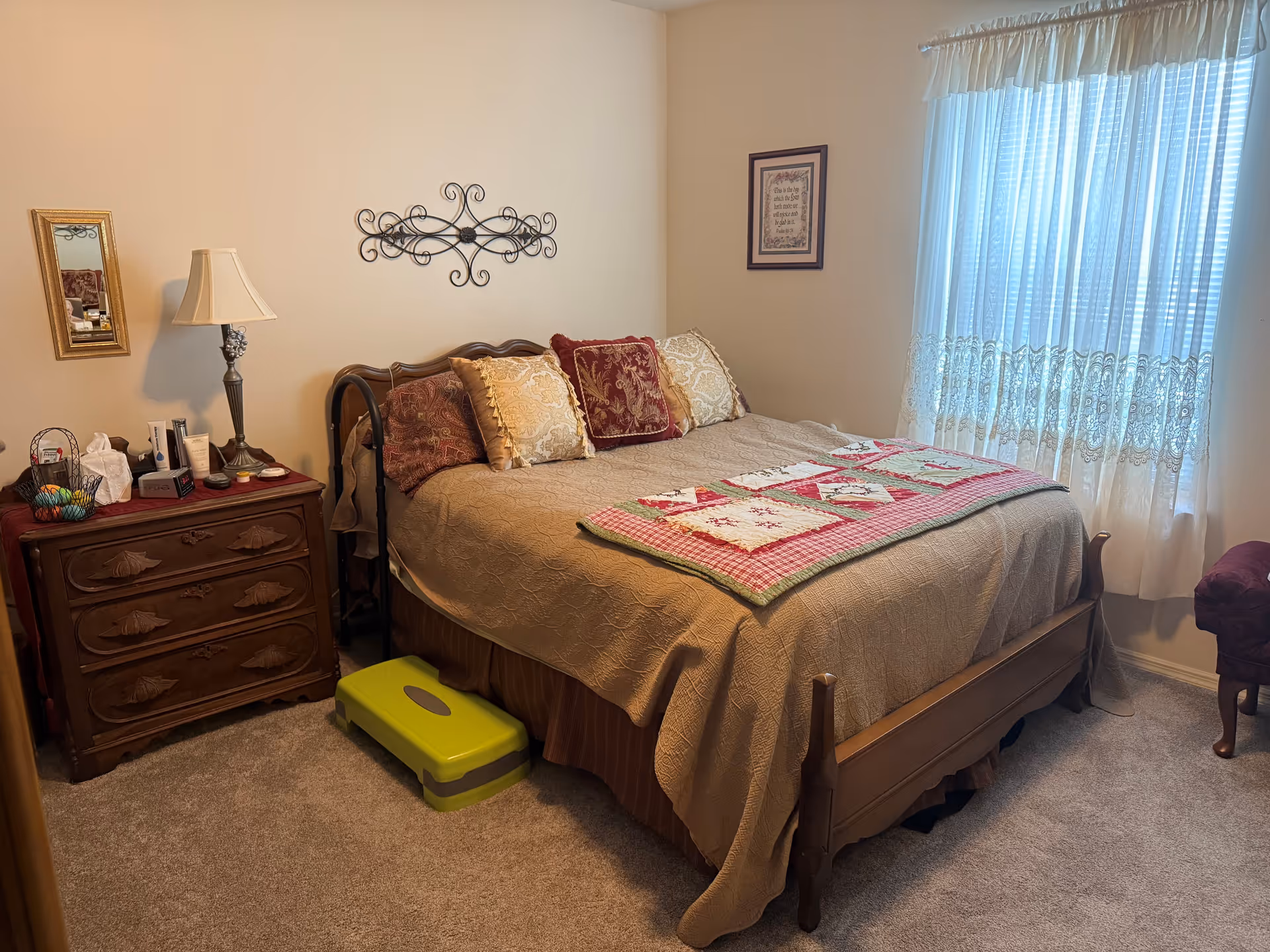A cozy bedroom with a wooden bed frame and a beige quilted bedspread. The bed is adorned with several decorative pillows and a colorful patchwork quilt at the foot. To the left of the bed is a wooden nightstand with a lamp, mirror, and various personal items. A green step stool is placed in front of the bed. A window with sheer white curtains allows natural light to fill the room. The walls are decorated with a metal wall art piece above the bed and a framed picture on the adjacent wall.
