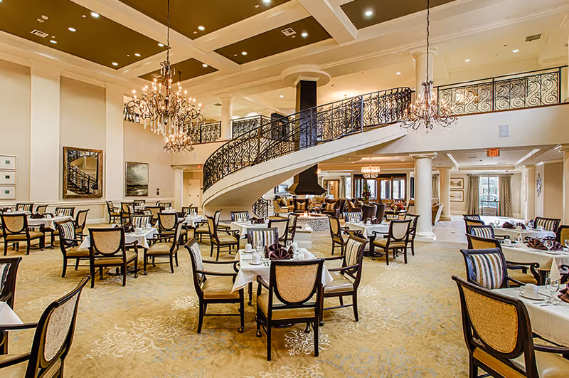 Elegant dining room in The Westmore Senior Living featuring multiple tables with chairs, white tablecloths, and place settings. The room has a grand curved staircase with ornate black railings, chandeliers hanging from the ceiling, and large windows allowing natural light.