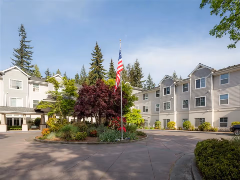 Exterior view of Cogir of Mill Creek senior living facility showing a circular driveway with landscaped center island featuring various trees and plants, including a prominent American flag on a flagpole. The building is three stories tall with multiple windows and light-colored siding, surrounded by tall evergreen trees under a clear blue sky.