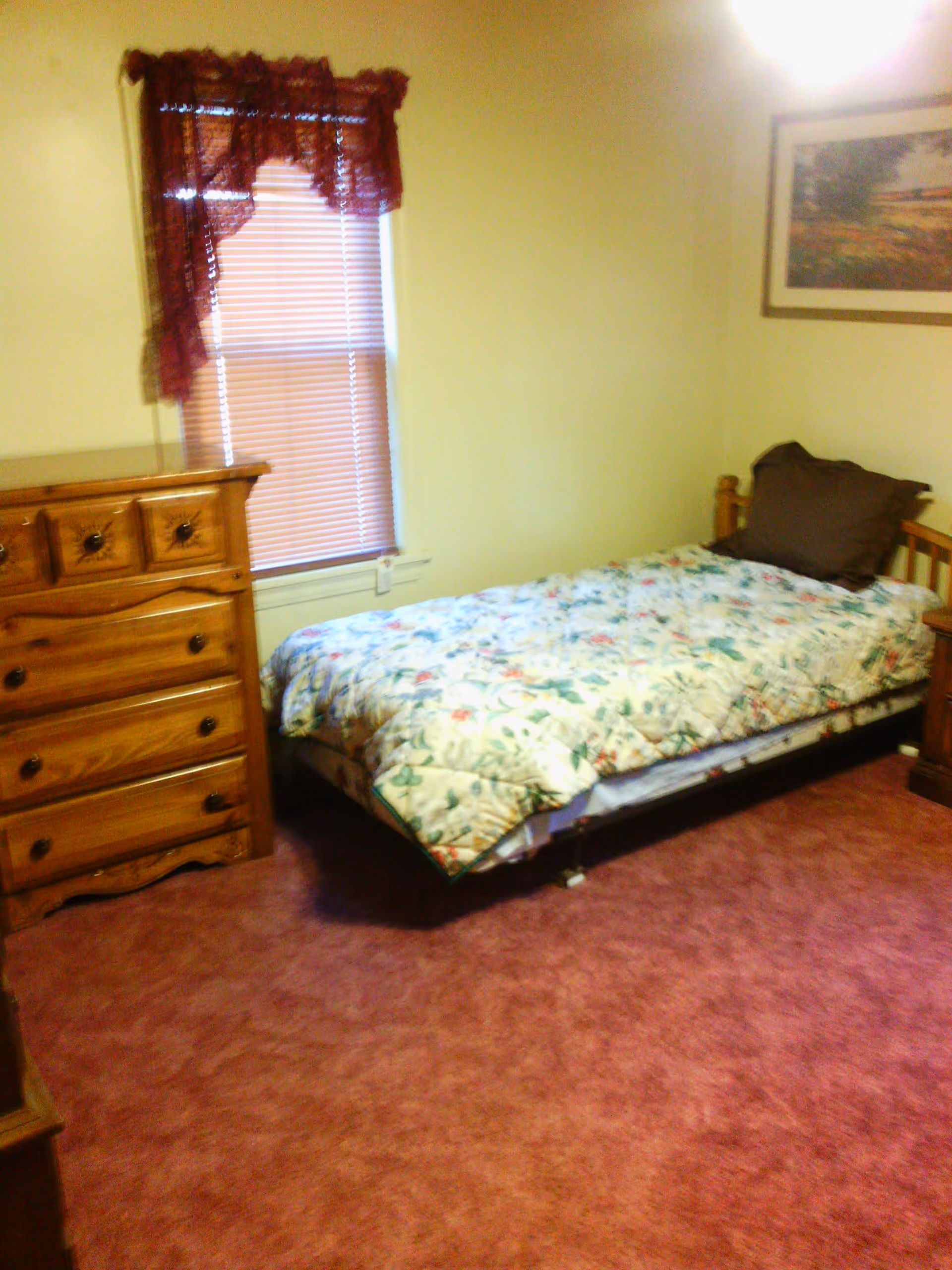 Small bedroom with a single bed covered in a floral comforter, wooden dresser, and a window with blinds and a burgundy valance.