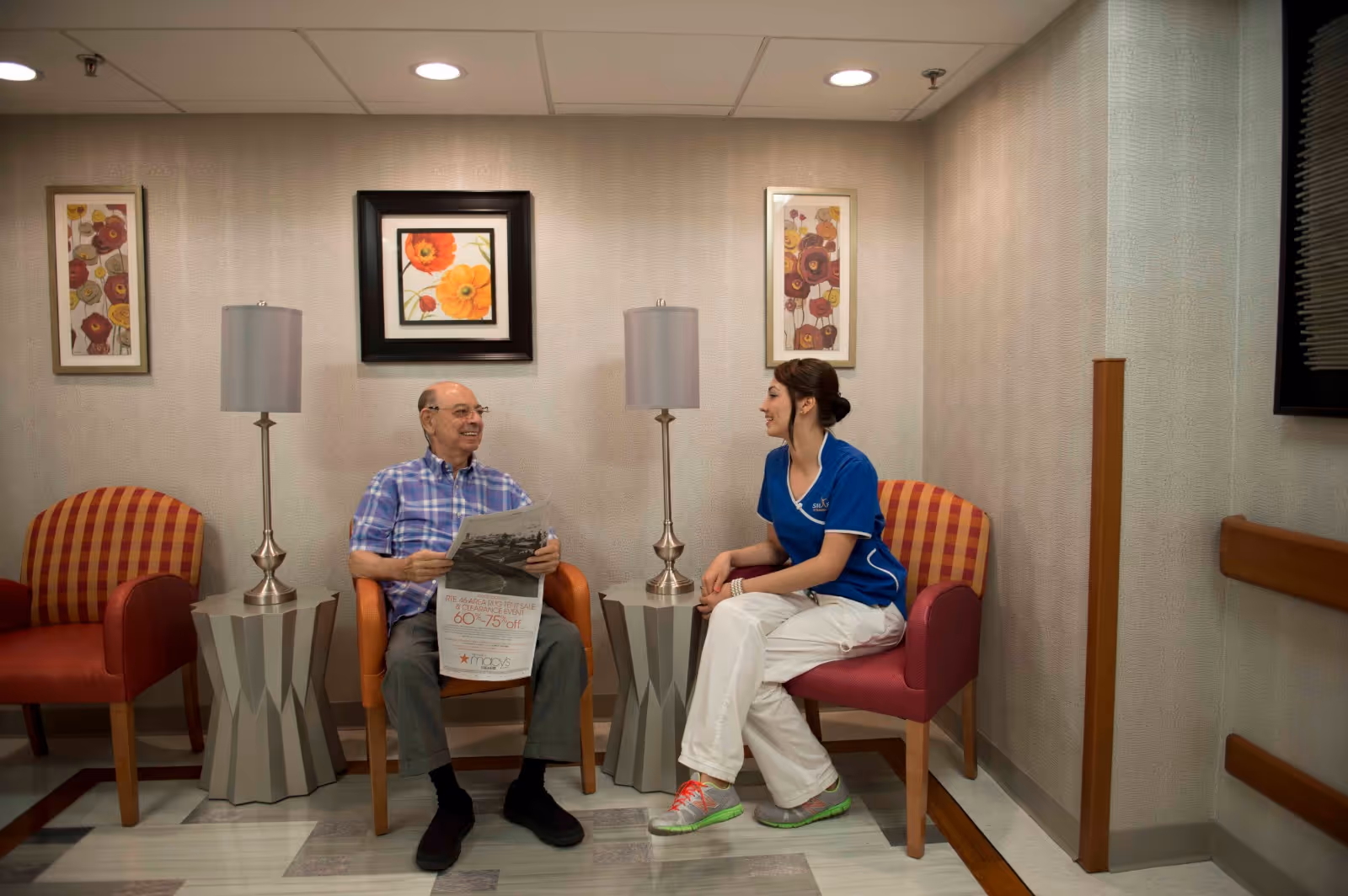 An elderly man and a female healthcare worker sitting and talking in a waiting area. The man is holding a newspaper and wearing a blue plaid shirt, while the woman is dressed in a blue medical uniform with white pants and colorful sneakers. The room has patterned chairs, two modern table lamps on side tables, and floral artwork on the wall behind them.