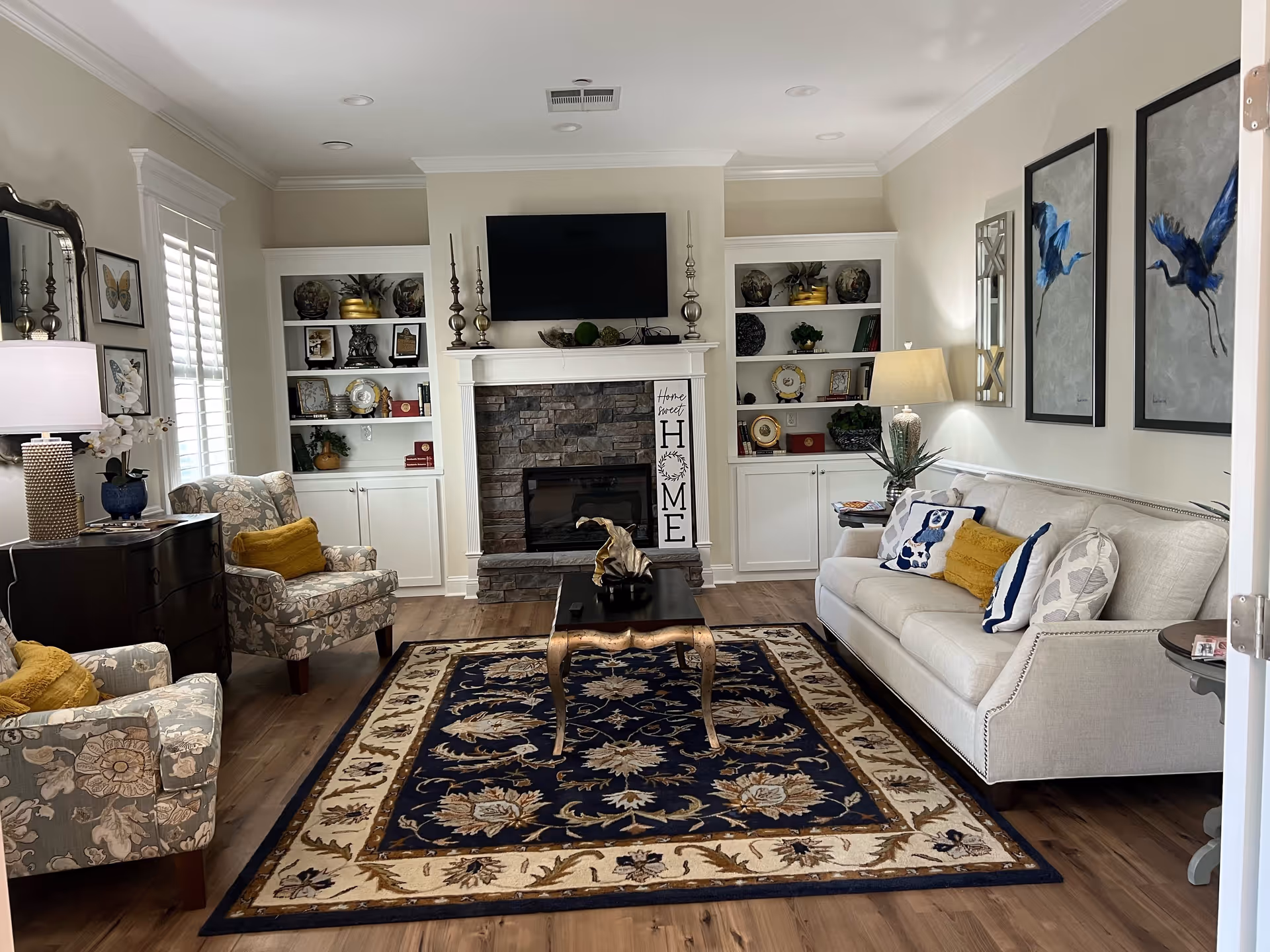 A cozy living room with a beige sofa adorned with decorative pillows, two floral patterned armchairs with yellow cushions, a dark wooden coffee table on a navy blue and beige patterned rug, built-in white shelves flanking a stone fireplace with a mounted flat-screen TV above it, and various decorative items including lamps, framed artwork, and plants.