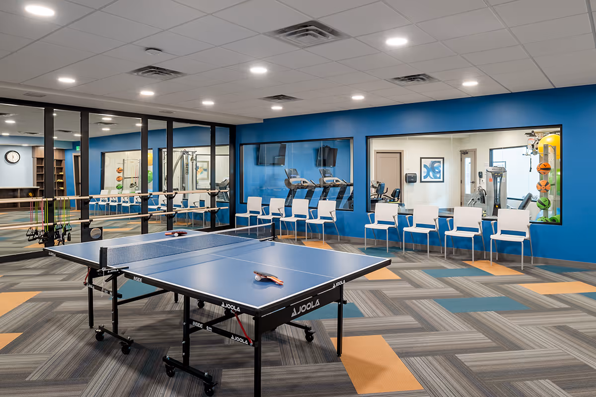 A recreational room with a blue JOOLA ping pong table in the center, surrounded by white chairs lined up against blue walls. The room has large windows looking into a gym area with exercise equipment and colorful exercise balls. The floor has a patterned carpet with gray, blue, and orange squares, and the ceiling has recessed lighting.