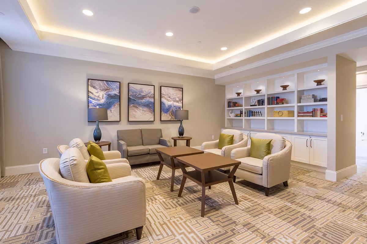 A well-lit living room area with a beige and brown patterned carpet, two white armchairs with green cushions, a gray loveseat, two wooden coffee tables, two side tables with blue lamps, and a built-in white bookshelf filled with books and decorative items. Three abstract paintings hang on the wall above the loveseat.