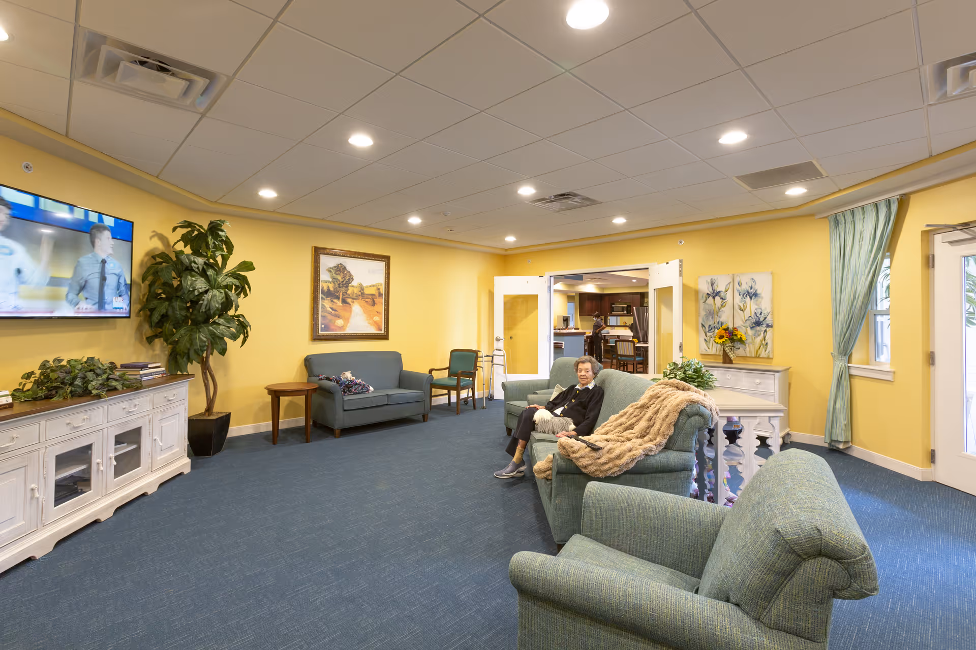 A cozy living room area in a senior living facility with yellow walls and blue carpet. The room features green upholstered sofas and chairs, a white wooden cabinet with plants and books, a large potted plant, and framed artwork on the walls. A television is mounted on the wall showing a program. An elderly woman is sitting on one of the sofas with a beige blanket draped over the armrest. Double doors lead to another room with a kitchen visible in the background.