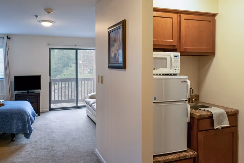 Interior view of a senior living facility room at Deer Park Assisted Living showing a small kitchenette with a microwave, mini fridge, sink, and wooden cabinets on the right. On the left, there is a bedroom area with a bed covered in a blue blanket, a TV on a stand, a white couch, and a sliding glass door leading to a balcony with a view of trees outside.