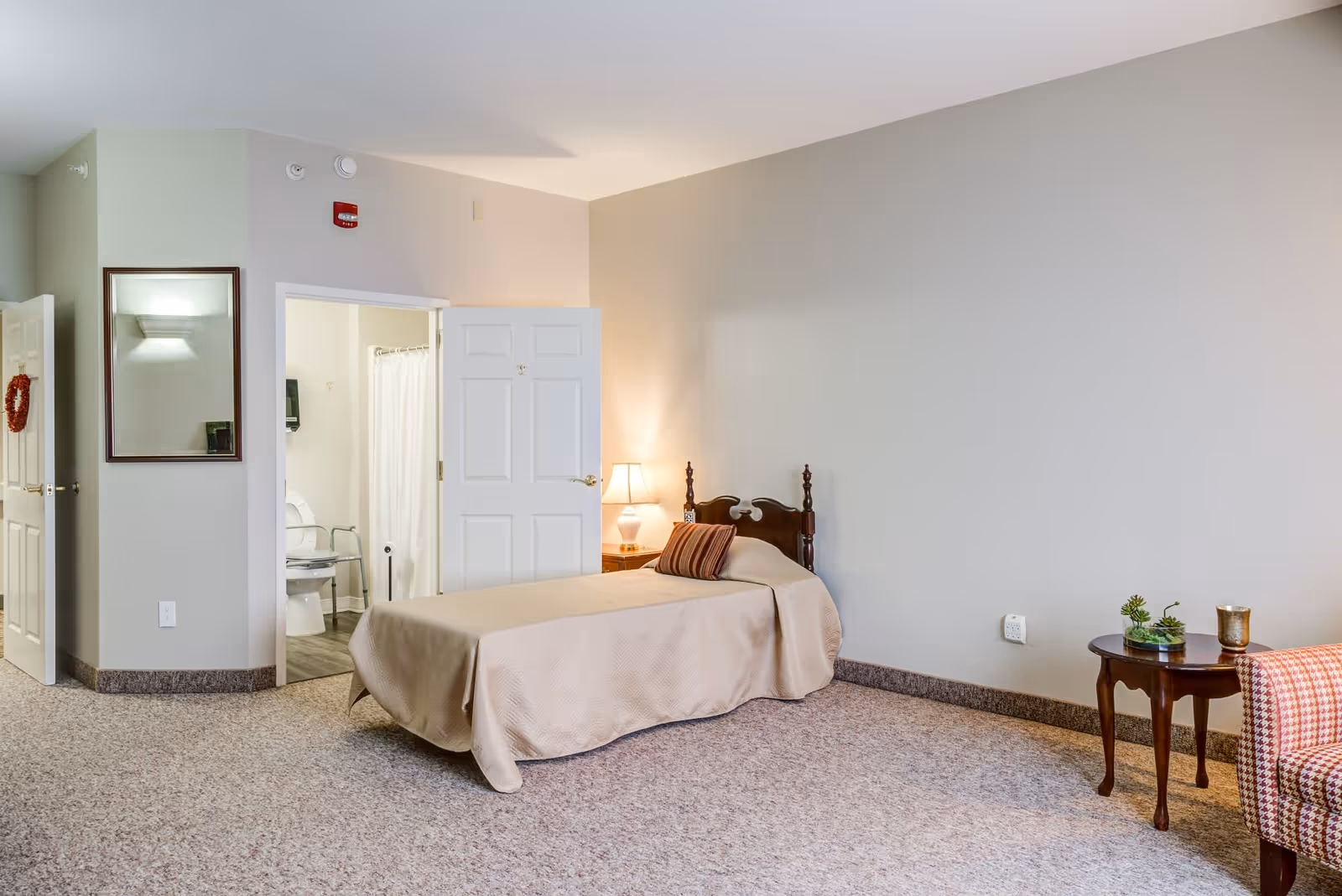 A senior living facility bedroom with a single bed covered in a beige bedspread and a striped pillow. There is a wooden nightstand with a lamp next to the bed. An open door reveals a bathroom with a toilet, grab bars, and a shower curtain. The room has beige walls, carpeted floor, a small round wooden table with a plant and a candle, and a patterned armchair partially visible on the right.