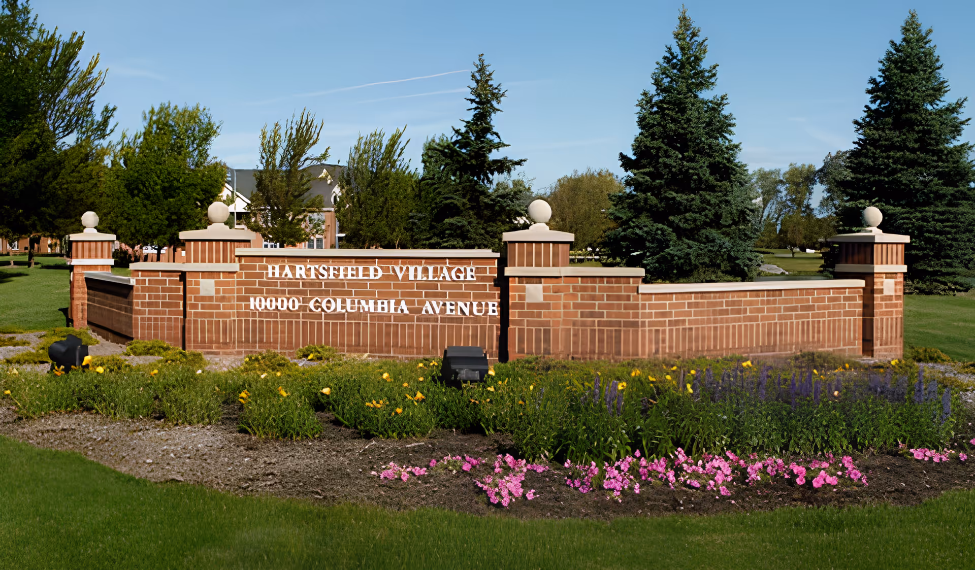 Brick entrance sign for Hartsfield Village with the address 10000 Columbia Avenue, surrounded by green grass, colorful flower beds, and tall evergreen trees under a clear blue sky.