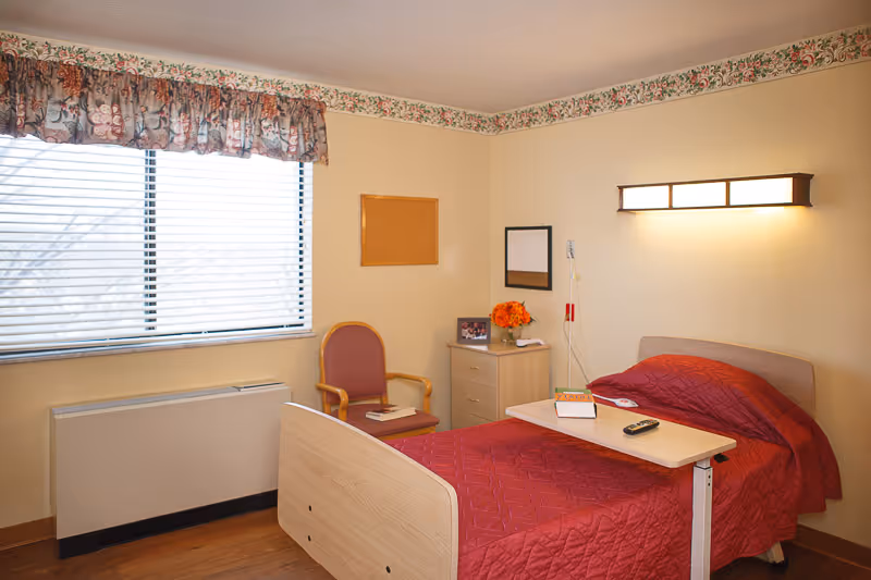 A small, cozy bedroom in a senior living facility with a single bed covered in a red quilt. A wooden overbed table holds a book and a remote control. Next to the bed is a light wood nightstand with a vase of orange flowers and a framed photo. A wooden chair with a red cushion sits by the window, which has blinds and a floral valance. The walls are light yellow with a floral border near the ceiling, and a rectangular wall light is mounted above the bed.