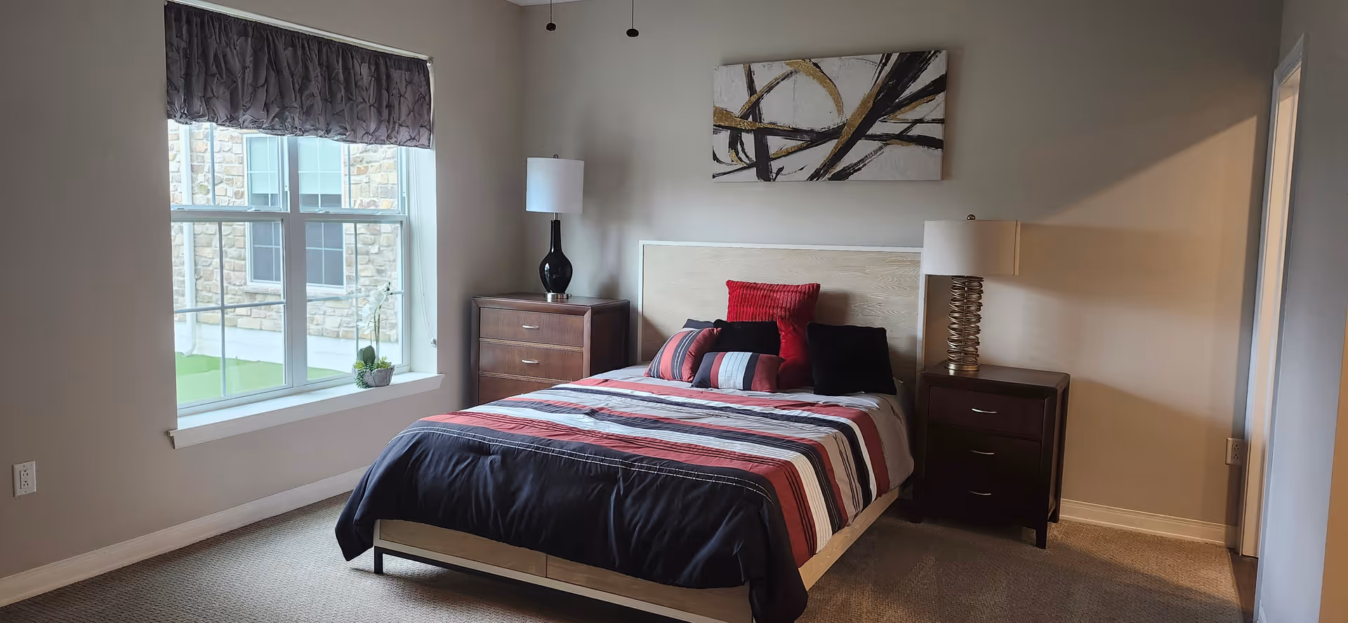 A bedroom with a bed featuring a striped red, black, and white comforter and several pillows. There are two dark wooden nightstands on either side of the bed, each with a lamp. A window with a dark valance is on the left wall, and a modern abstract painting hangs above the bed. The room has beige walls and carpeted floor.