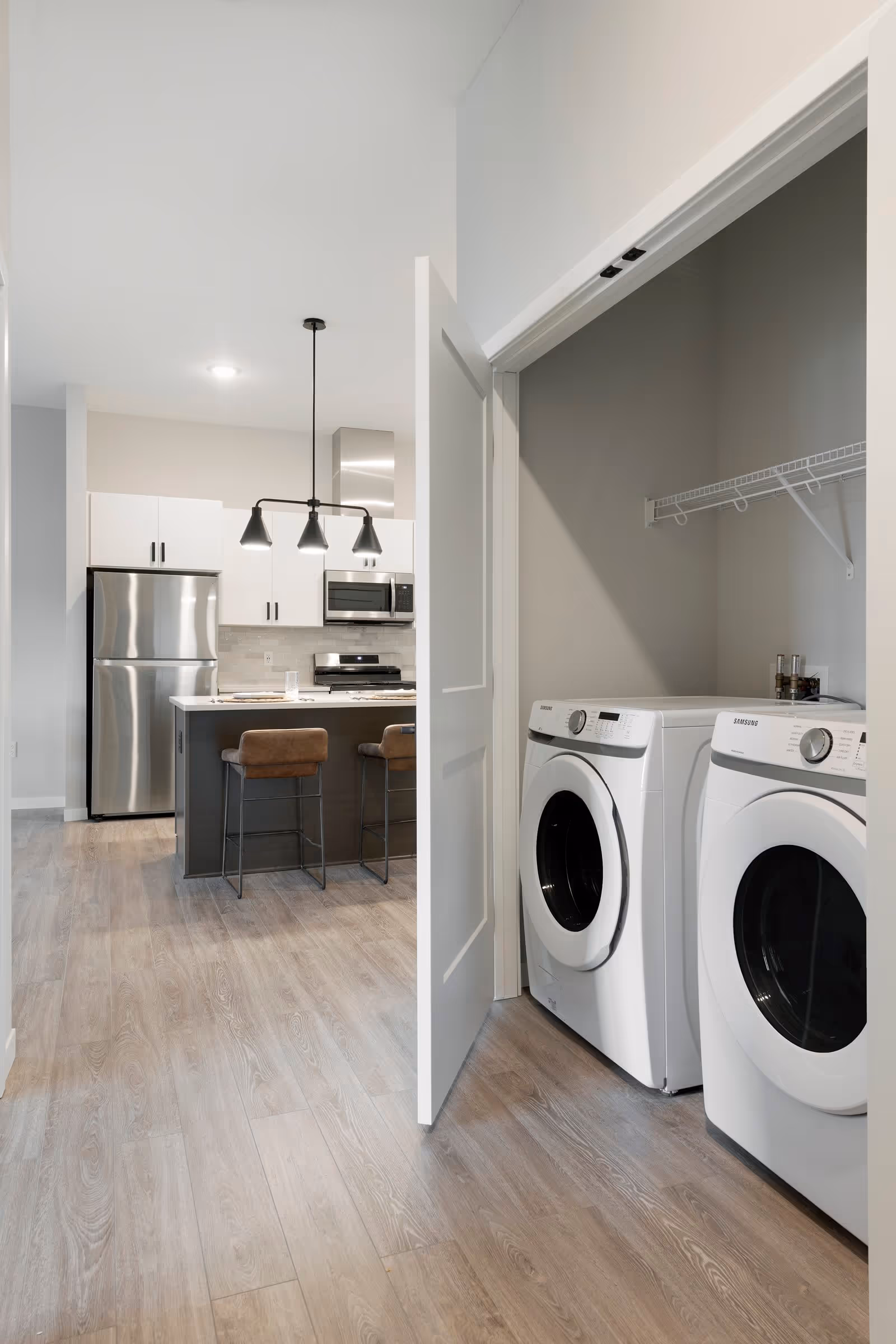 Modern kitchen with stainless steel refrigerator, stove, microwave, and a kitchen island with two brown bar stools. To the right, there is a laundry closet with a white front-loading washer and dryer, and a wire shelf above them.