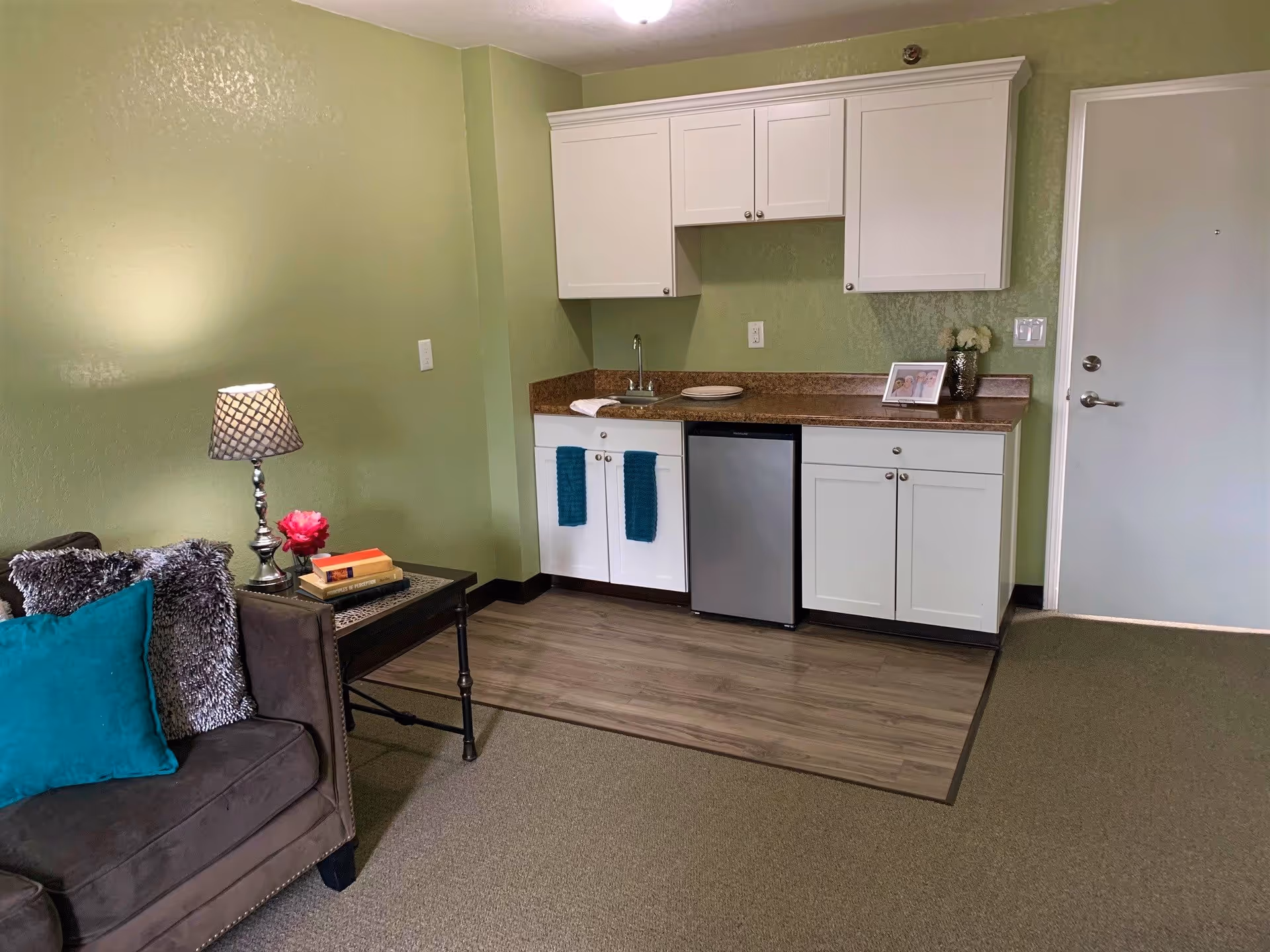 A small kitchenette area with white cabinets, a mini refrigerator, and a countertop with a sink. Next to the kitchenette is a brown couch with teal and gray pillows, a side table with a lamp, books, and a flower. The walls are painted light green, and there is a closed white door to the right.