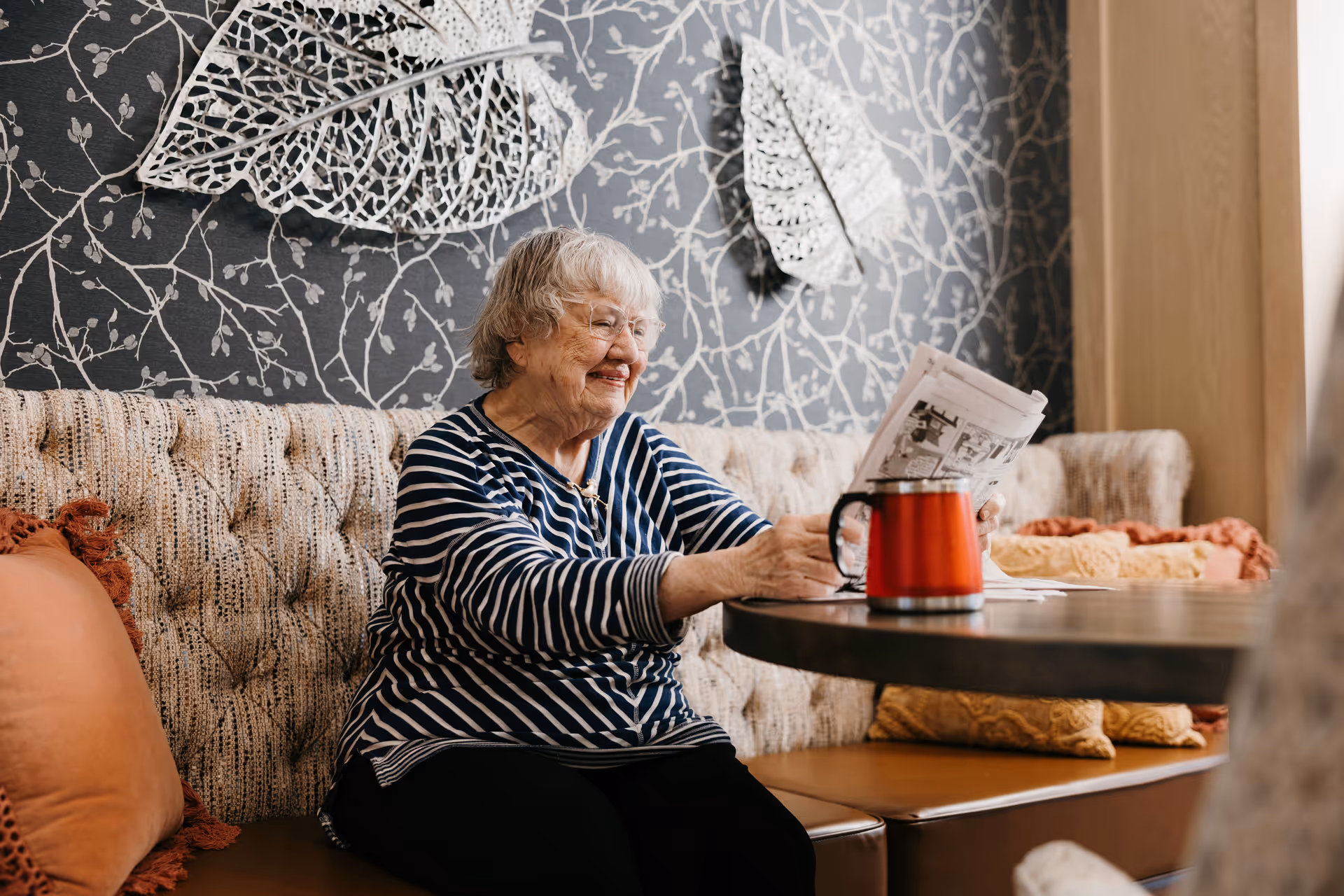 An elderly woman with gray hair and glasses sits on a cushioned bench in a cozy room, smiling as she reads a newspaper. The wall behind her features decorative metal leaf art on a patterned wallpaper. A red mug is on the round table in front of her, and there are several pillows on the bench.