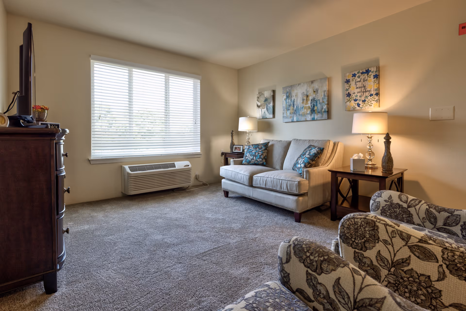 A cozy living room with a beige couch adorned with two patterned blue pillows, two floral armchairs, a wooden side table with a lamp and decorative items, a large window with white blinds, and a wooden dresser with a TV on top. The walls are decorated with three pieces of artwork.