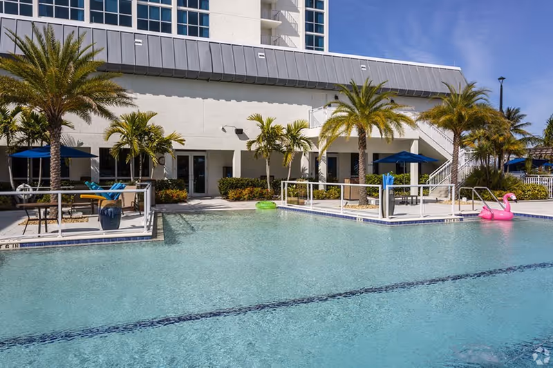 Outdoor swimming pool with palm trees, lounge chairs, umbrellas and a pink flamingo float in front of a multi-story building.