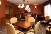 Warm communal dining and seating room with a square wooden table, upholstered chairs, a wall-mounted TV, and pendant lamps.