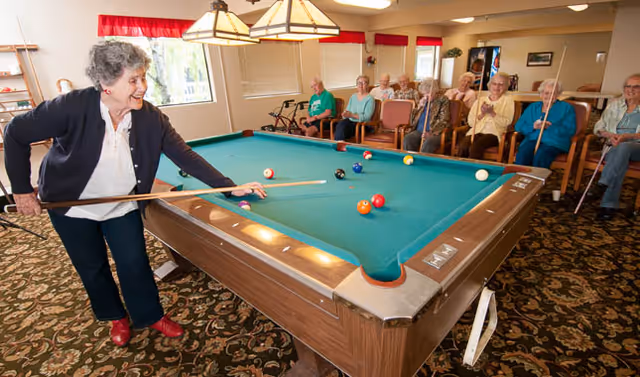 An elderly woman playing pool in a senior living facility game room while a group of elderly people sit in chairs watching and smiling.