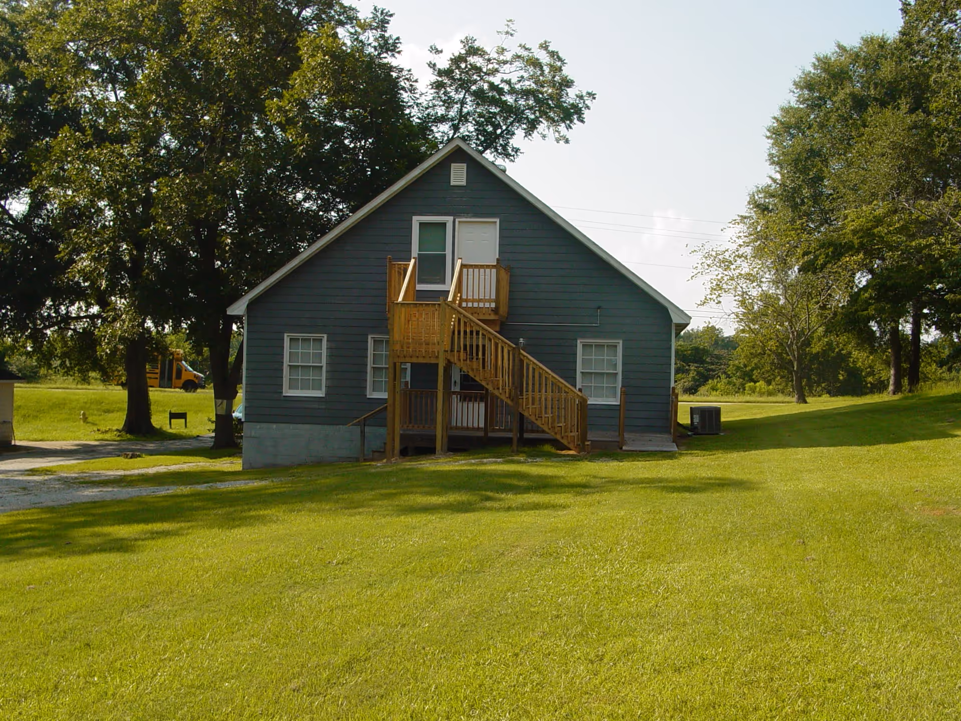 A small gray two-story house with white-framed windows and a wooden staircase leading to a second-floor door. The house is surrounded by a well-maintained green lawn and several large trees under a clear sky.