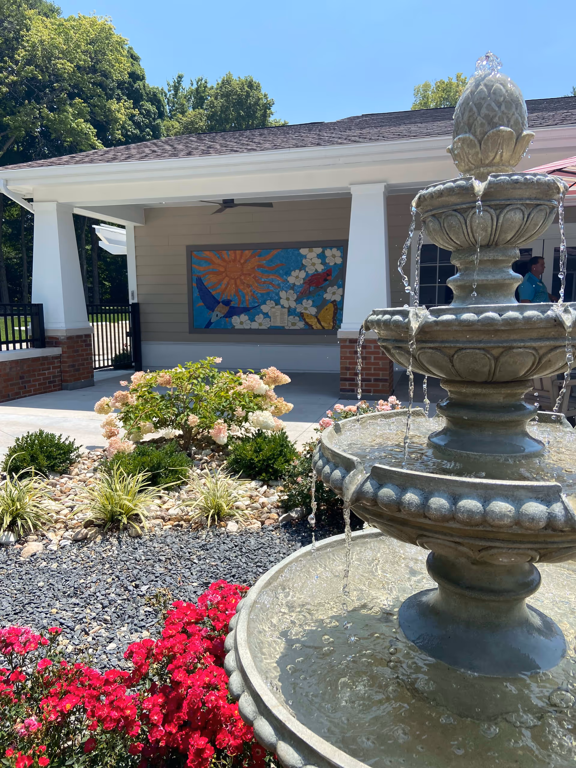 Stone tiered fountain with water flowing in front of a landscaped entrance featuring flowering bushes and a covered building facade with a colorful mural.