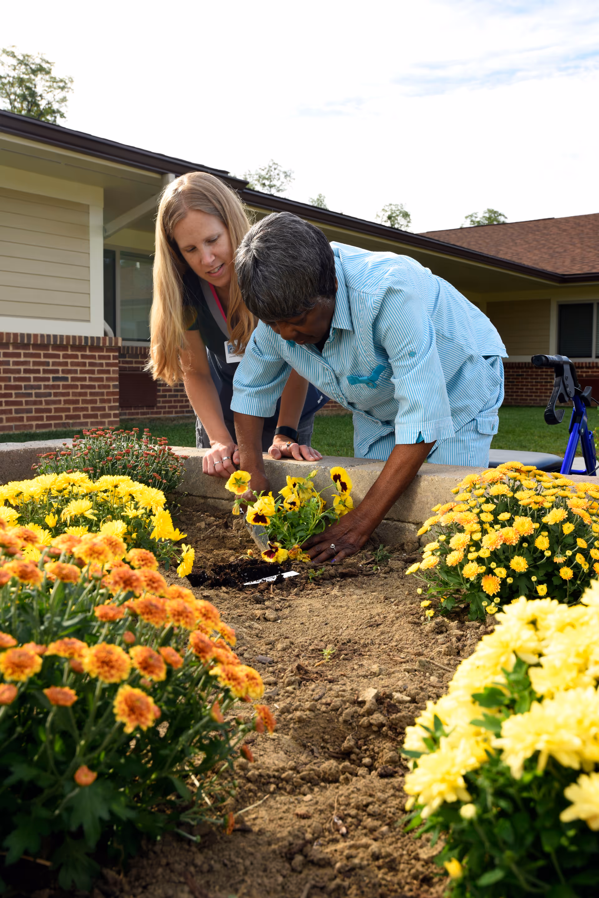 An elderly woman is planting yellow flowers in a garden bed outside a building, assisted by a caregiver who is watching and supporting her. The garden bed is surrounded by blooming yellow and orange flowers, and a walker is visible nearby.