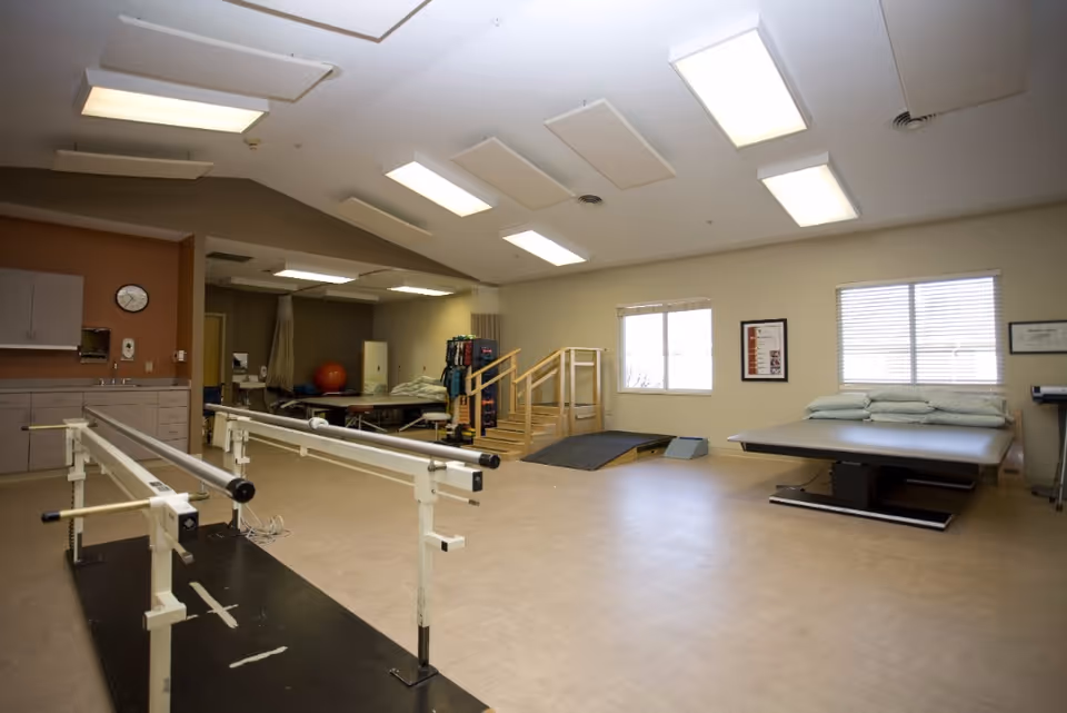 A spacious physical therapy room with parallel bars for walking exercises, a therapy table with pillows, a small set of wooden stairs with a ramp, exercise equipment, and bright overhead lighting. The room has beige walls, two windows with blinds, and a clock on the wall above cabinets and a sink.