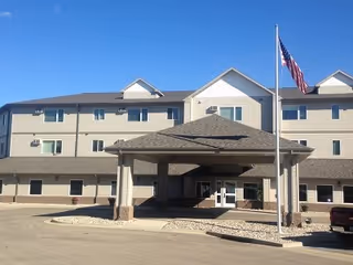 Exterior view of a three-story senior living facility building with beige siding and a covered entrance. An American flag is flying on a flagpole near the entrance under a clear blue sky.