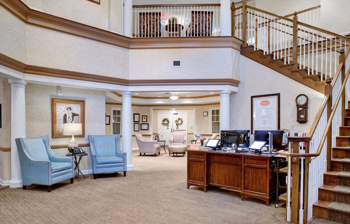 Interior view of a senior living facility lobby with a wooden reception desk equipped with computers, a staircase with wooden handrails and white balusters, light blue armchairs with a small table and lamp, framed artwork on the walls, and a seating area with armchairs and a round ottoman in the background.