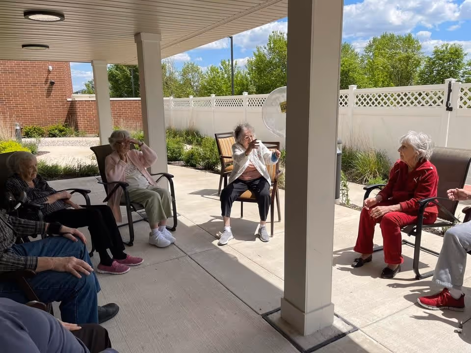 A group of elderly people sitting in chairs in a covered outdoor patio area, engaging in an activity with a large transparent ball. The setting includes a white fence, green bushes, and a partly cloudy sky in the background.