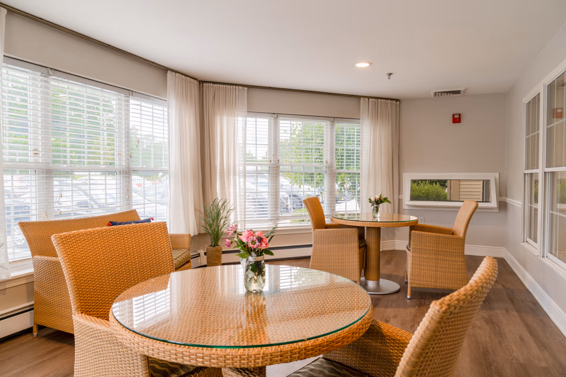 A bright and cozy sitting area with two round glass-top wicker tables, each surrounded by wicker chairs. There is a wicker loveseat with cushions near large windows with white blinds and sheer curtains. Small flower arrangements are placed on the tables, and a rectangular mirror is mounted on the wall. The room has light-colored walls and wood flooring.