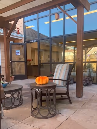 Covered courtyard patio with a cushioned chair and side tables—one holding a pumpkin—in front of large glass windows and a door.