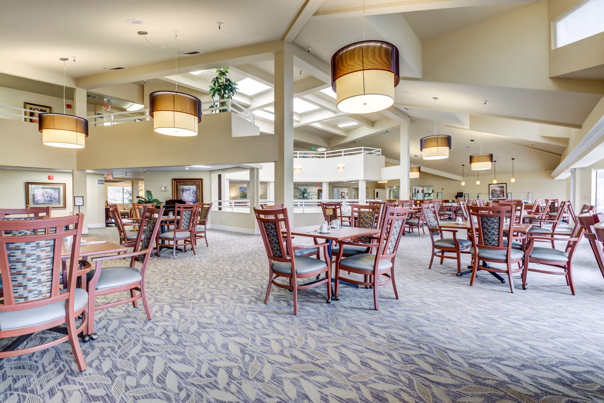 Spacious dining area in Deer Park Retirement Community with multiple wooden tables and chairs arranged neatly on a patterned carpet. The room features high ceilings with large hanging light fixtures and skylights, creating a bright and airy atmosphere. There are framed pictures on the walls and large windows allowing natural light to enter.