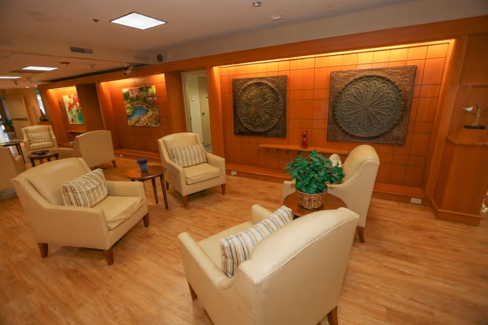 A cozy seating area in a senior living facility with beige armchairs and small wooden tables arranged on a wooden floor. The walls are decorated with framed artwork and two large ornamental wall plaques. There is a potted plant on one of the tables, and the lighting is warm and inviting.