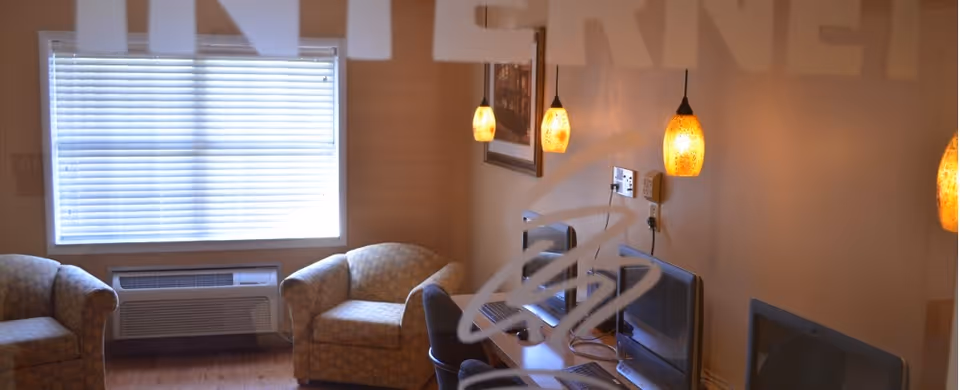 A small lounge with two upholstered armchairs, a window with blinds, pendant lights, and TVs on a desk visible through frosted glass.
