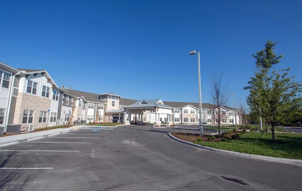 Exterior view of The Crossings at Riverview senior living facility showing a large two-story building with a covered entrance, parking spaces, landscaped greenery, and a clear blue sky.