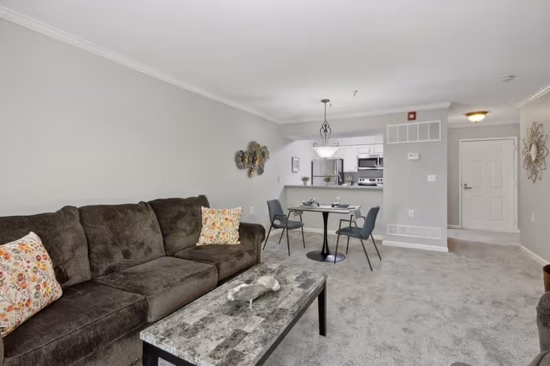 A living room and dining area in an apartment with a dark brown sofa adorned with floral patterned pillows, a rectangular marble coffee table, and a small round dining table set with two chairs and place settings. The background shows a kitchen with white cabinets and stainless steel appliances, and a hallway leading to a white door. The walls are painted light gray and the floor is carpeted.