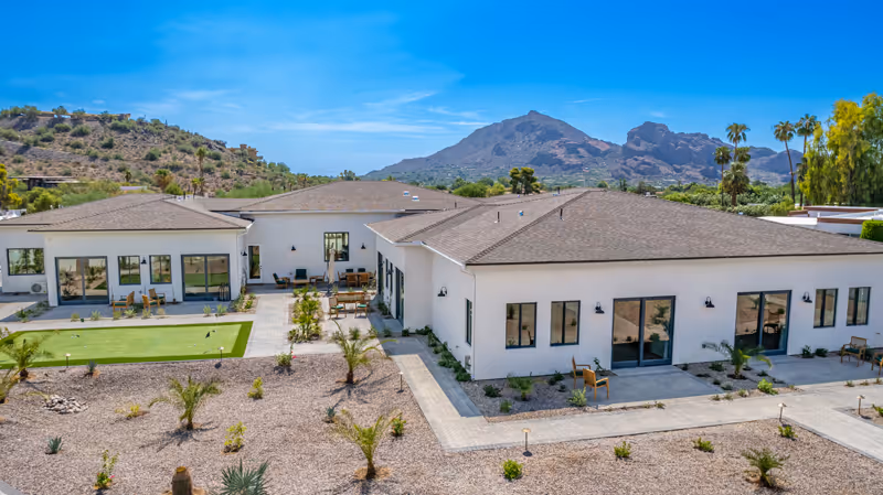 Exterior view of a single-story senior living facility building with white walls and multiple windows. The building is surrounded by a landscaped area with small palm trees, desert plants, and a paved walkway. In the background, there are mountains under a clear blue sky.