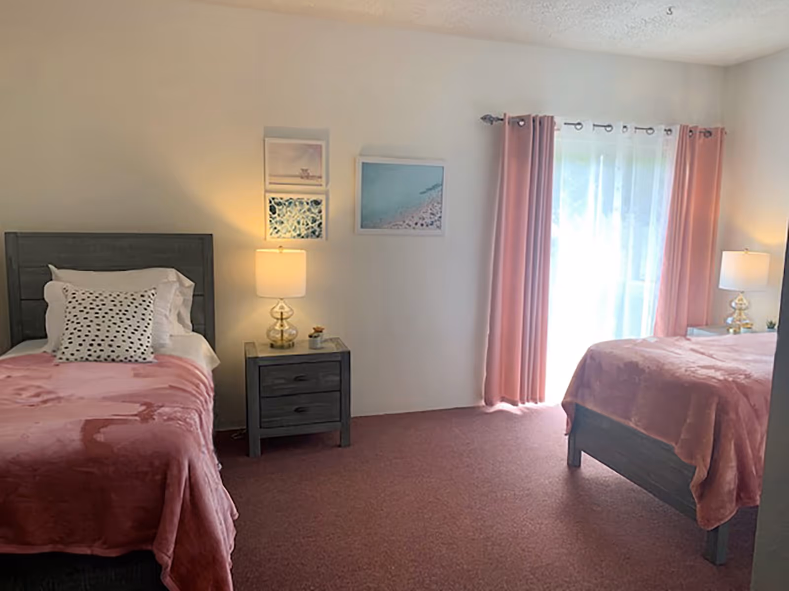A bedroom with two beds covered in pink blankets, each with white pillows. There is a nightstand with a lamp between the beds. The room has pink curtains and sheer white curtains covering a window or glass door. Three framed pictures hang on the wall above the nightstand.