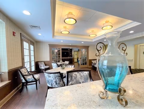 A well-lit interior room with multiple tables and chairs arranged for seating. The room features a coffered ceiling with recessed lighting and hanging light fixtures. A large decorative blue glass vase with metal scrollwork is placed on a marble countertop in the foreground. The walls have a light patterned wallpaper, and there are French doors and windows letting in natural light.